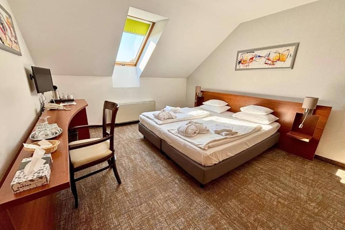 A hotel room with a bed, a desk with a computer, a chair, a window, and artwork on the walls.