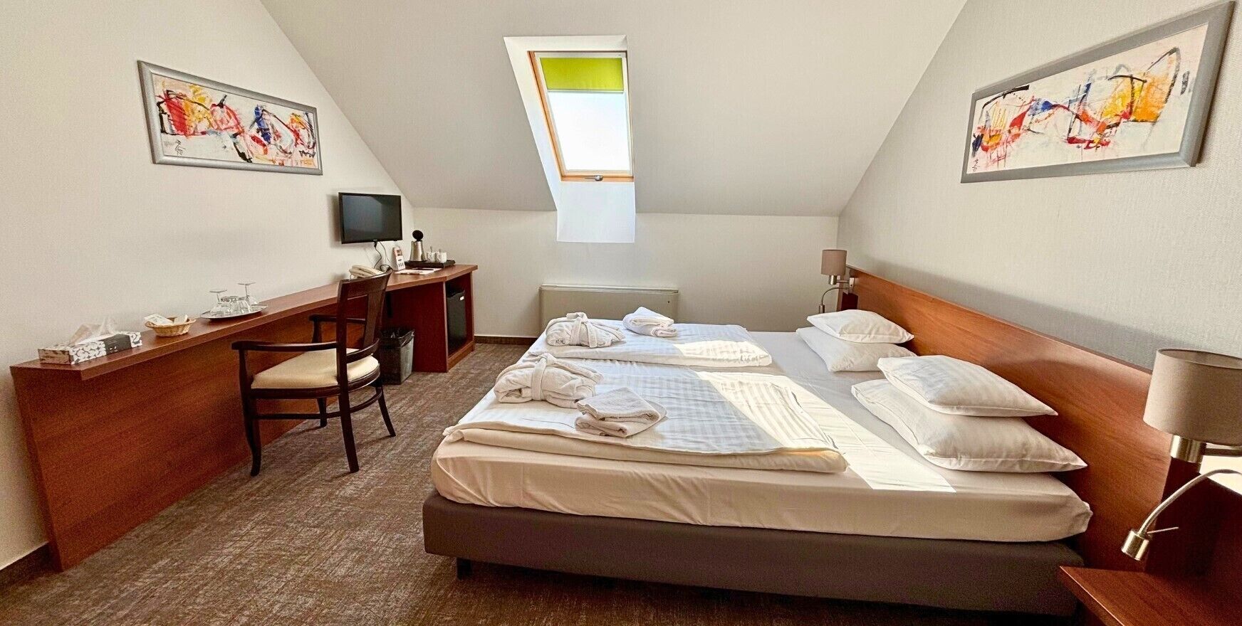 A hotel room with a bed, desk, chair, and artwork on the walls.