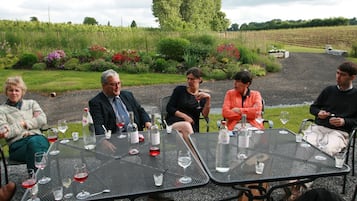 A group of people seated at a table outdoors with wine glasses and bottles, surrounded by a garden with various plants and flowers.
