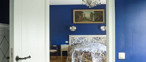 A bedroom with a bed, a chair, a nightstand, a painting on the wall, and a chandelier.