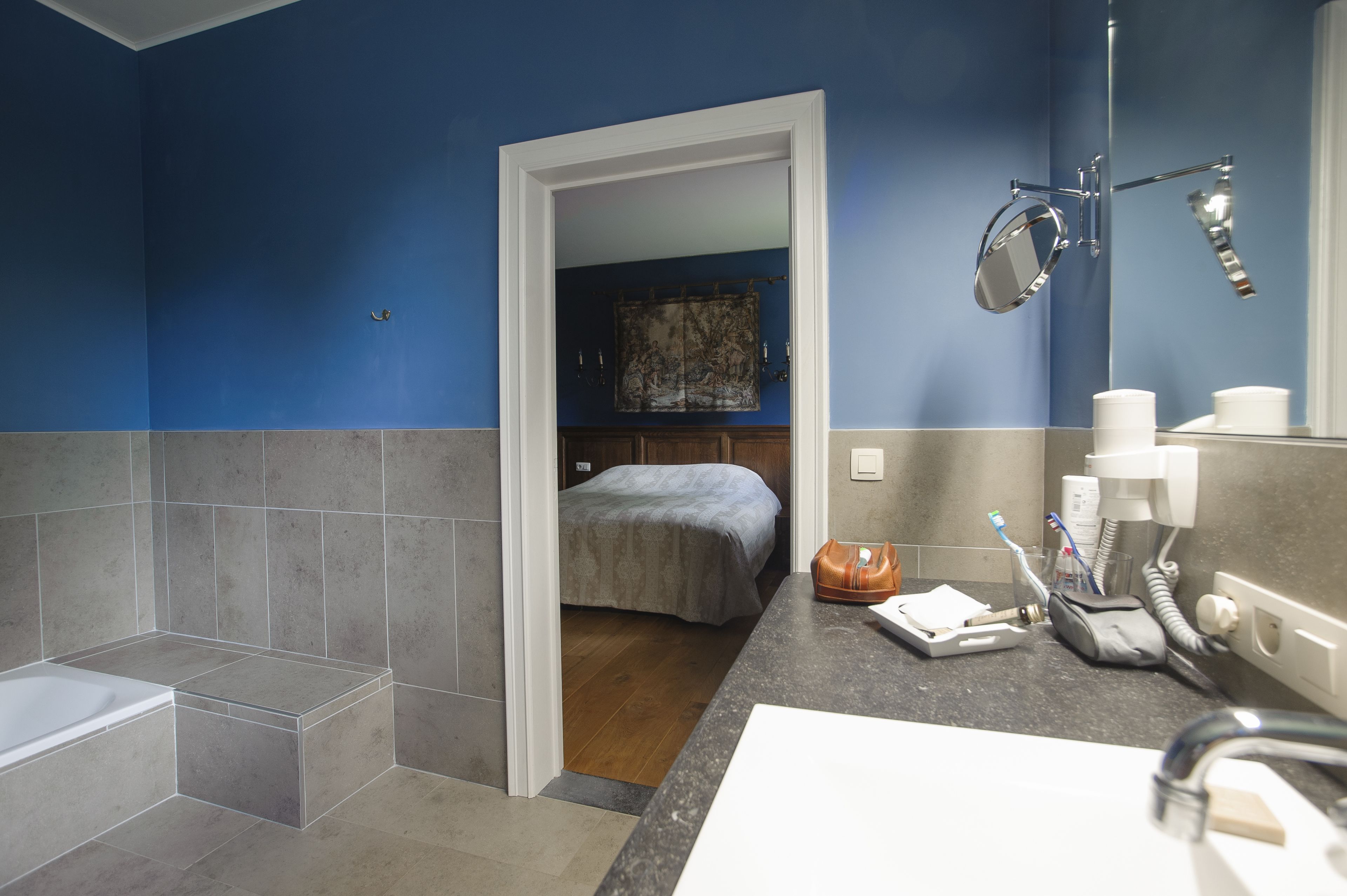 A bathroom with a bathtub, a shower, a sink with a mirror, and a toilet. There is a bed visible through an open door in the background.