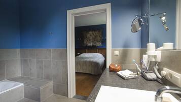 A bathroom with a bathtub, a shower, a sink with a mirror, and a toilet. There is a bed visible through an open door in the background.