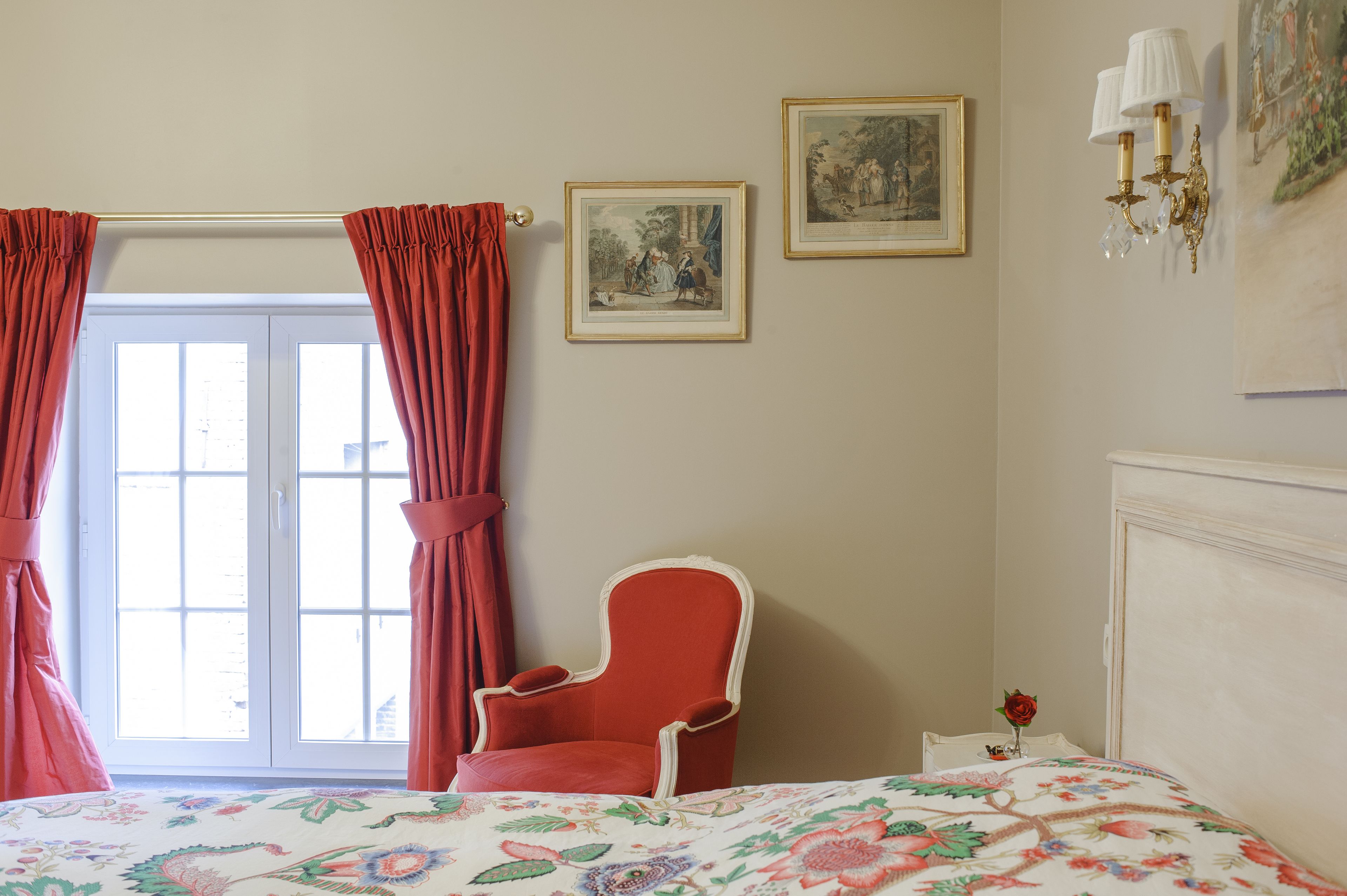 Un dormitorio con cama, una silla, una ventana con cortinas y cuadros en la pared.