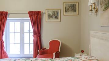Un dormitorio con cama, una silla, una ventana con cortinas y cuadros en la pared.