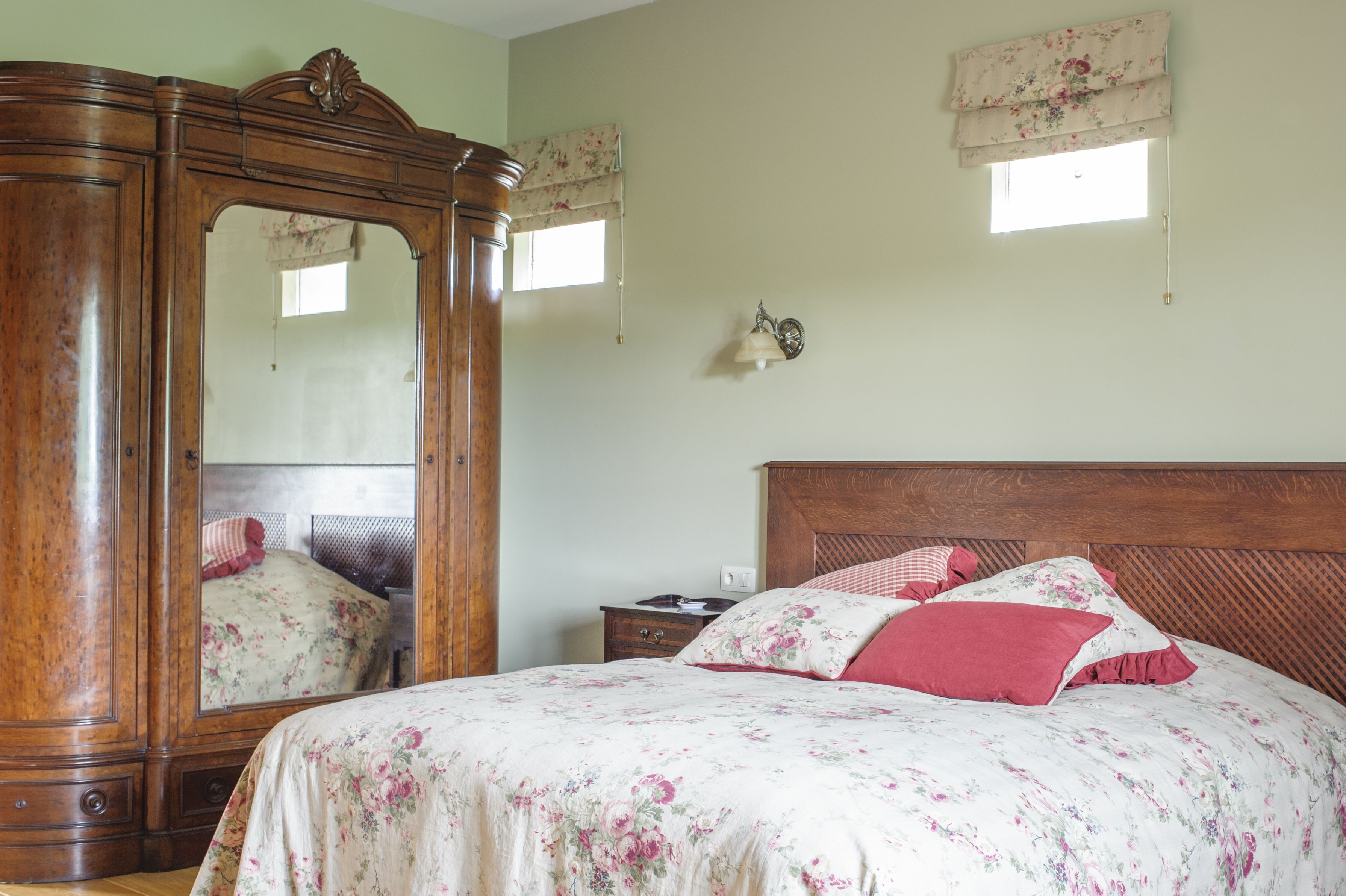 A bedroom with a wooden wardrobe, a bed with floral bedding, a nightstand, and a wall-mounted lamp.
