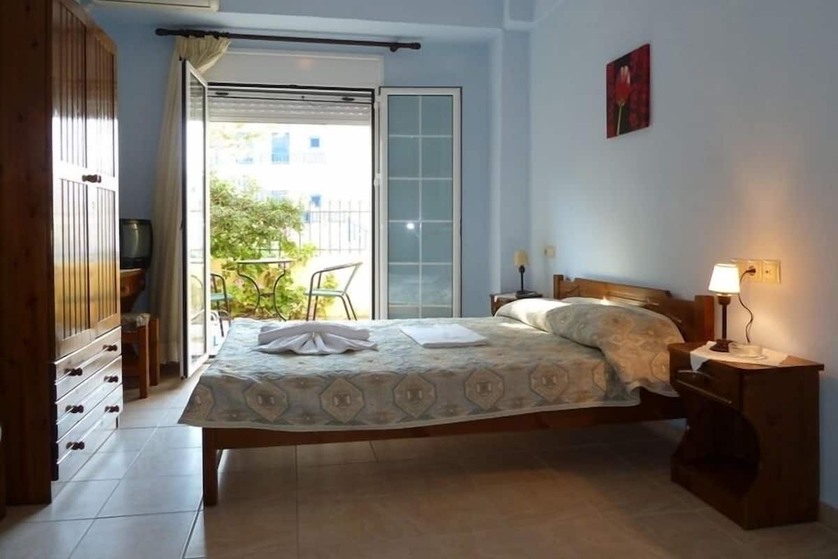 A bedroom with a bed, a nightstand, a wardrobe, a sliding glass door leading to a balcony, and a small table with chairs outside.