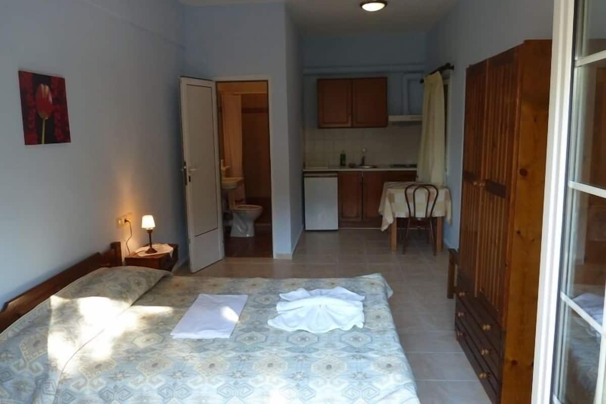 A compact room with a bed, a bedside table with a lamp, a small kitchenette, and a bathroom visible through an open door.