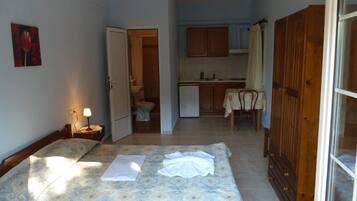 Habitación pequeña con cama, mesita de noche con lámpara, kitchenette y baño visible a través de una puerta abierta.