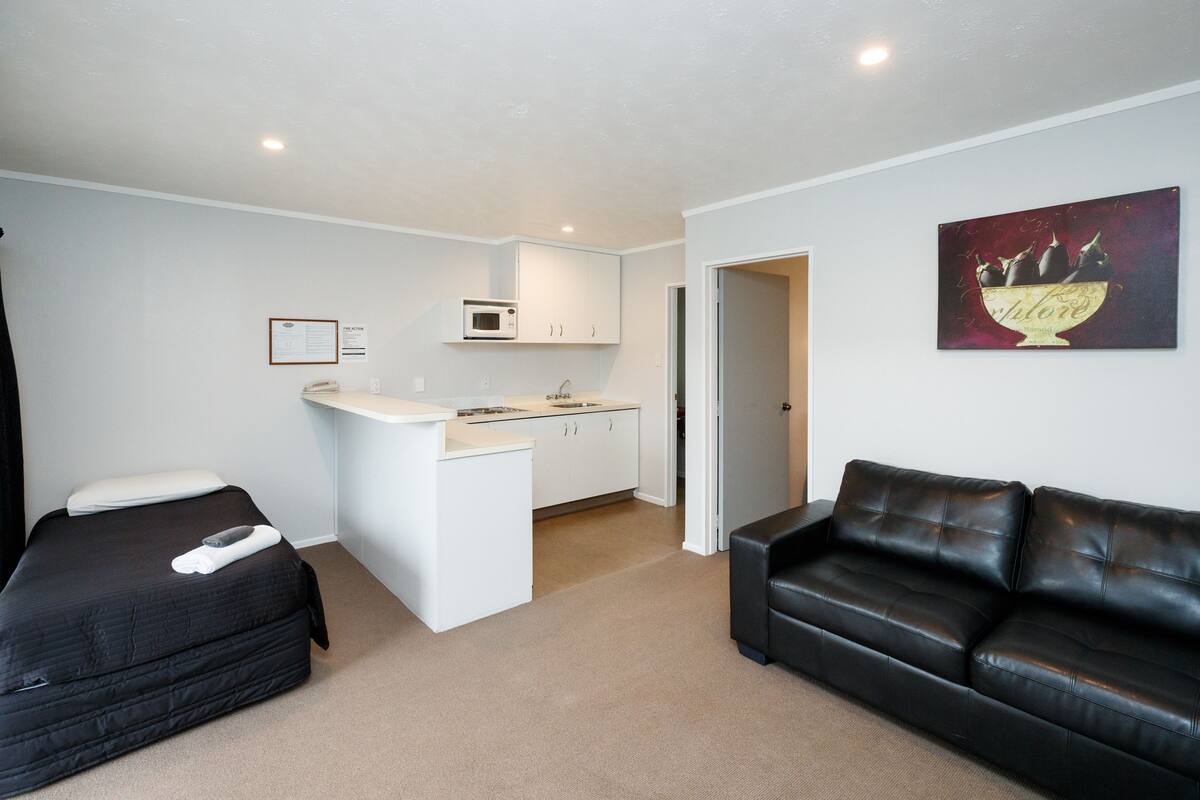 A compact living space with a kitchenette, a black leather sofa, and a bed with a pillow and remote control.