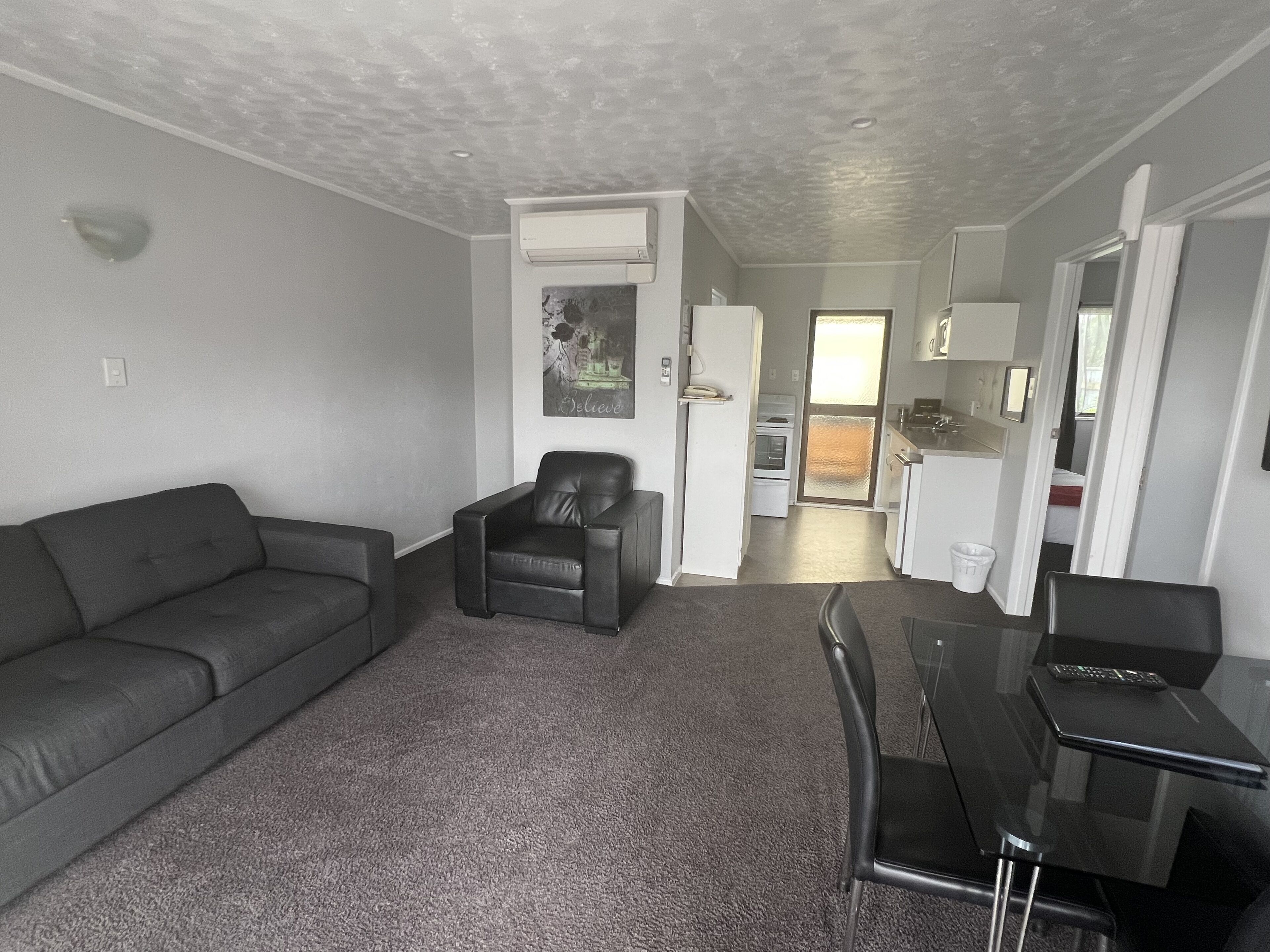 A compact living space with a grey sofa, a black leather chair, a dining table with chairs, a kitchen area with a fridge, and a small oven.