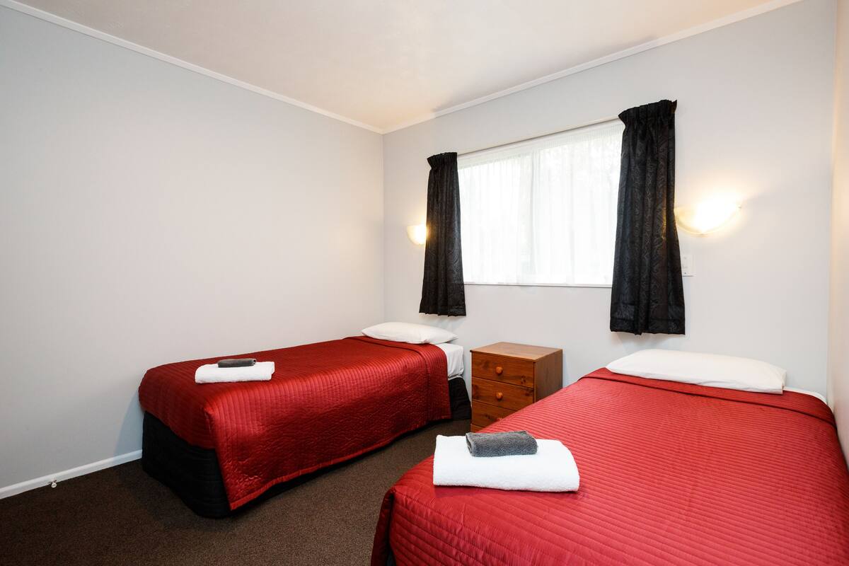 A hotel room with two single beds, a wooden nightstand, a small dresser, and a window with curtains.