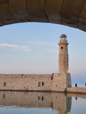 A stone lighthouse with a circular base and a dome on top, situated next to a stone wall with a water body in front. - Casa Maistra Residence (Rethymno)