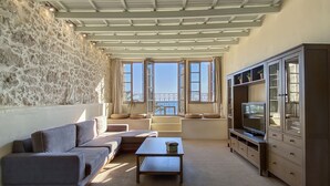A living room with a stone wall, a sofa, a coffee table, a TV on a wooden cabinet, and large windows with a view. - Casa Maistra Residence (Rethymno)