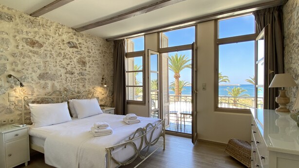 A bedroom with a stone wall, a bed with white linens, a nightstand, a dresser, and a balcony with a view of the sea. - Casa Maistra Residence (Rethymno)