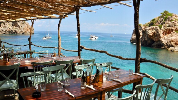 Un restaurant en bord de mer, avec des tables et des chaises en bois, offrant une vue sur une côte rocheuse et la mer.