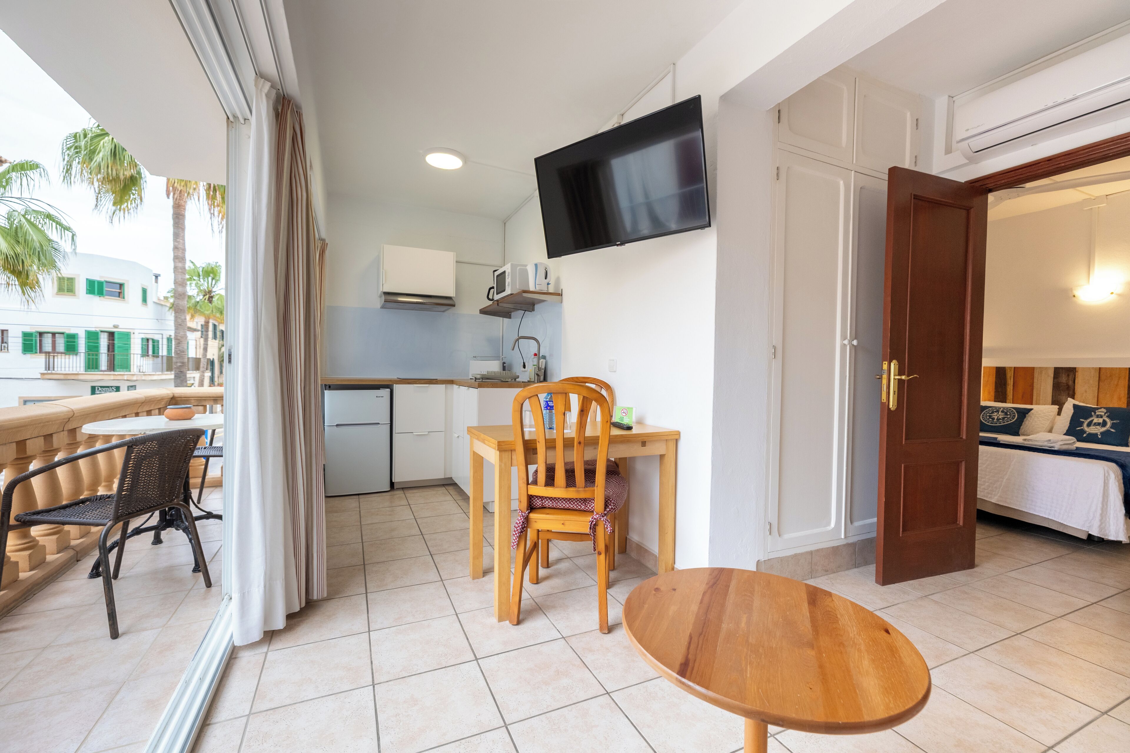 A compact hotel room with a balcony, a dining area, a TV mounted on the wall, and a bed with a view.