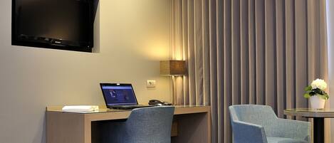 A modern hotel room with a wooden desk, two blue chairs, a flat-screen TV, a lamp, and a potted plant.