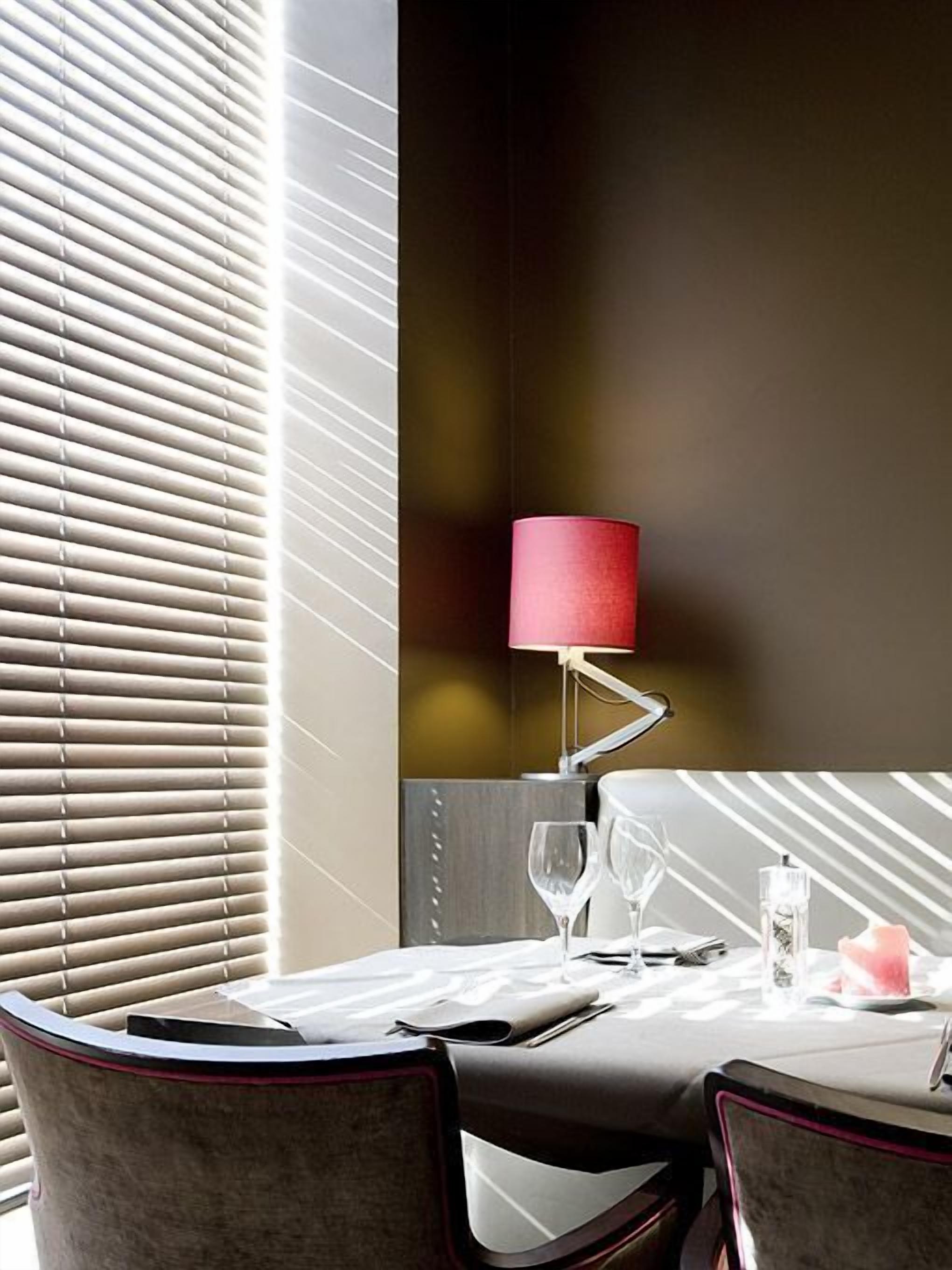Un espace de salle à manger moderne avec une table dressée pour deux personnes, une lampe rouge et une vue à travers des stores.