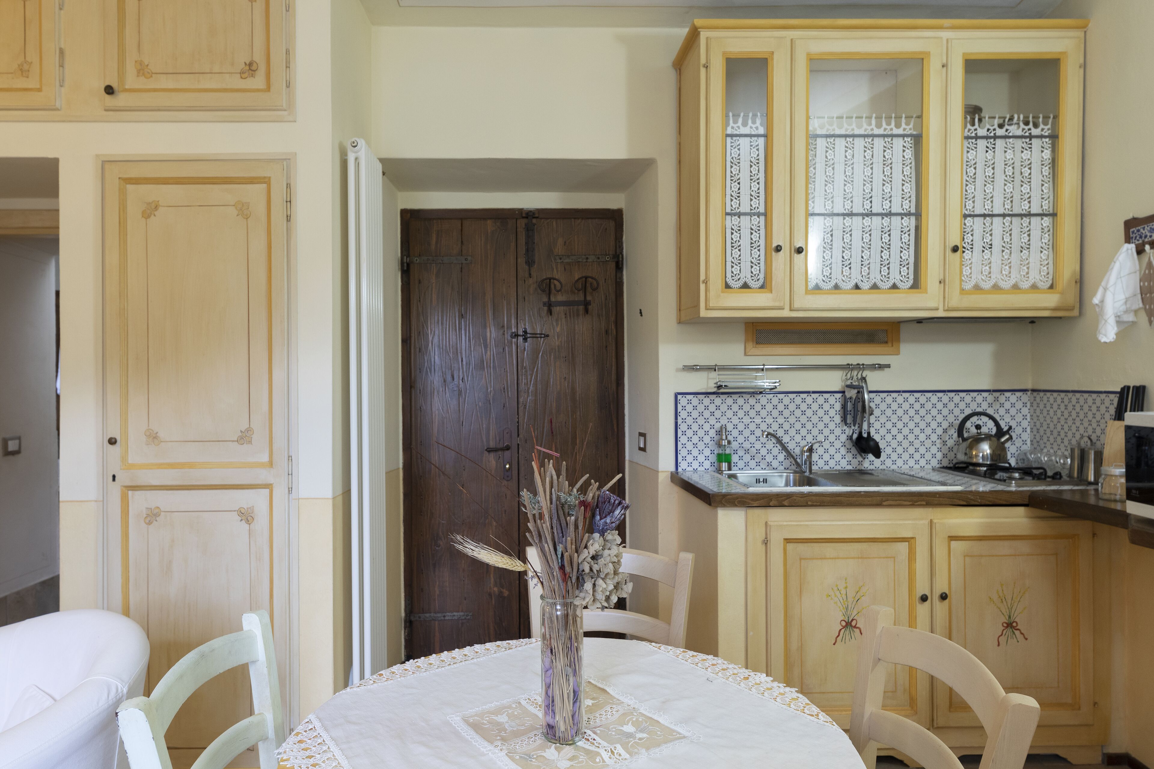 A kitchen with wooden cabinets, a white table with a lace tablecloth, and a vase with dried flowers.