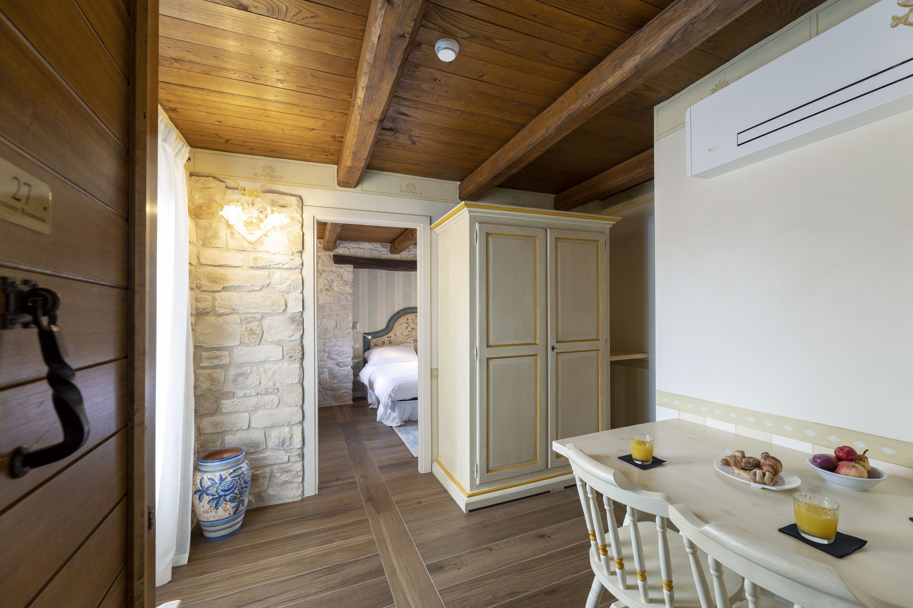 A wooden interior with a stone wall, a bed, a wardrobe, and a breakfast table with fruit and juice.