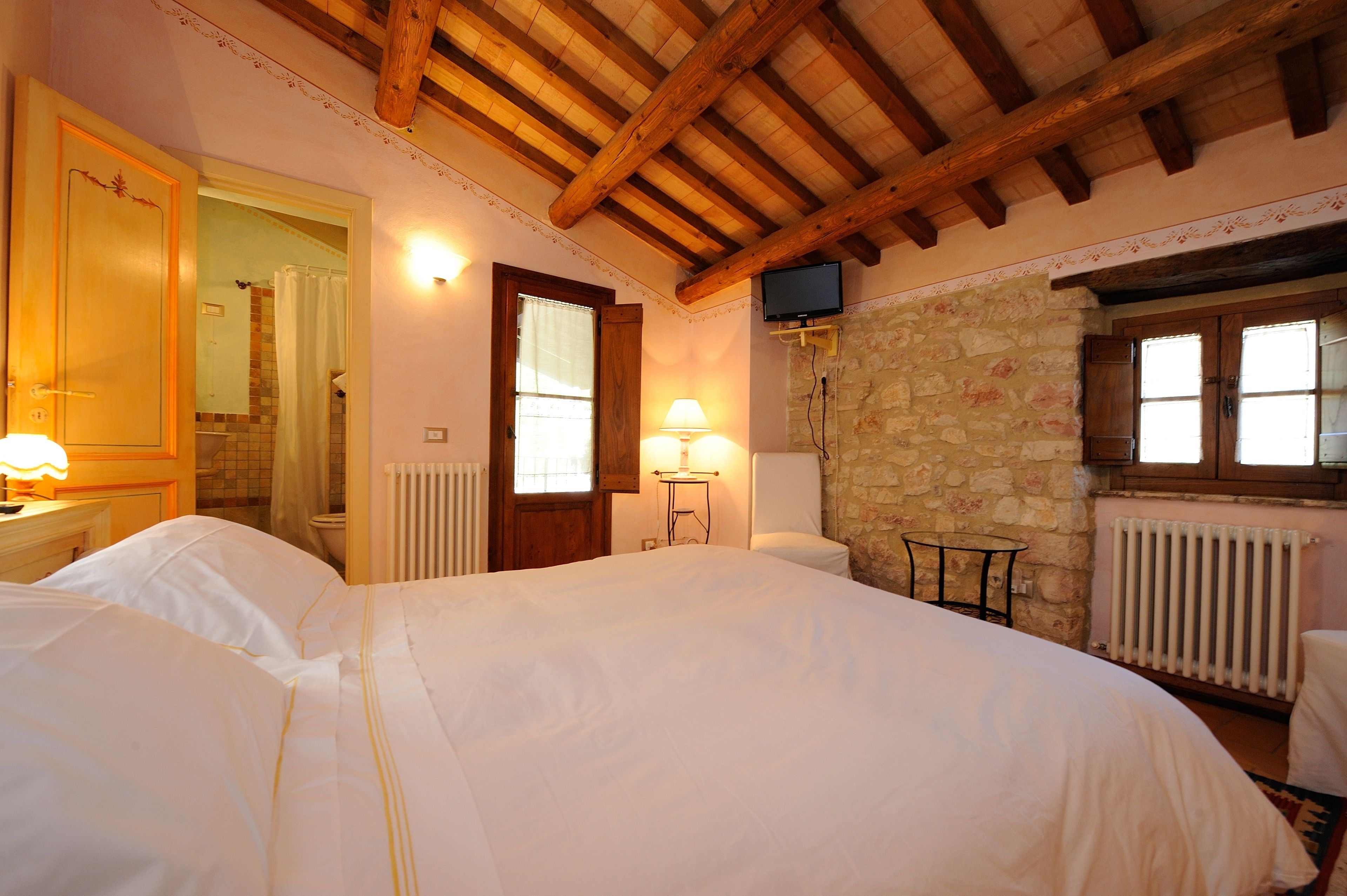 A bedroom with a large bed, wooden ceiling beams, a stone wall, a small table, and a television mounted on the wall.