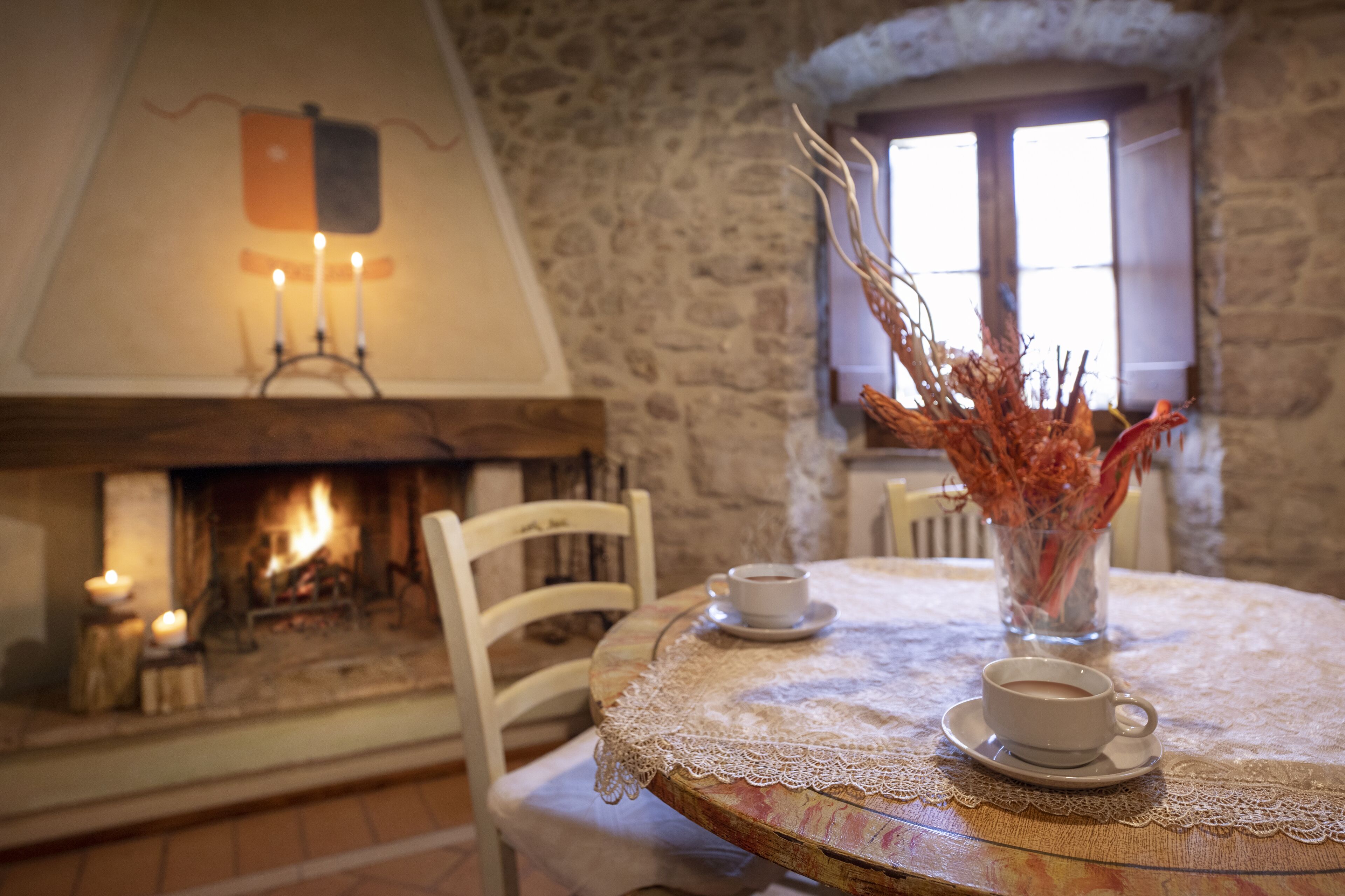 Un coin repas accueillant avec une table ronde, des chaises blanches et un vase contenant des branches séchées. On aperçoit une cheminée avec des bougies et une fenêtre avec des rideaux.