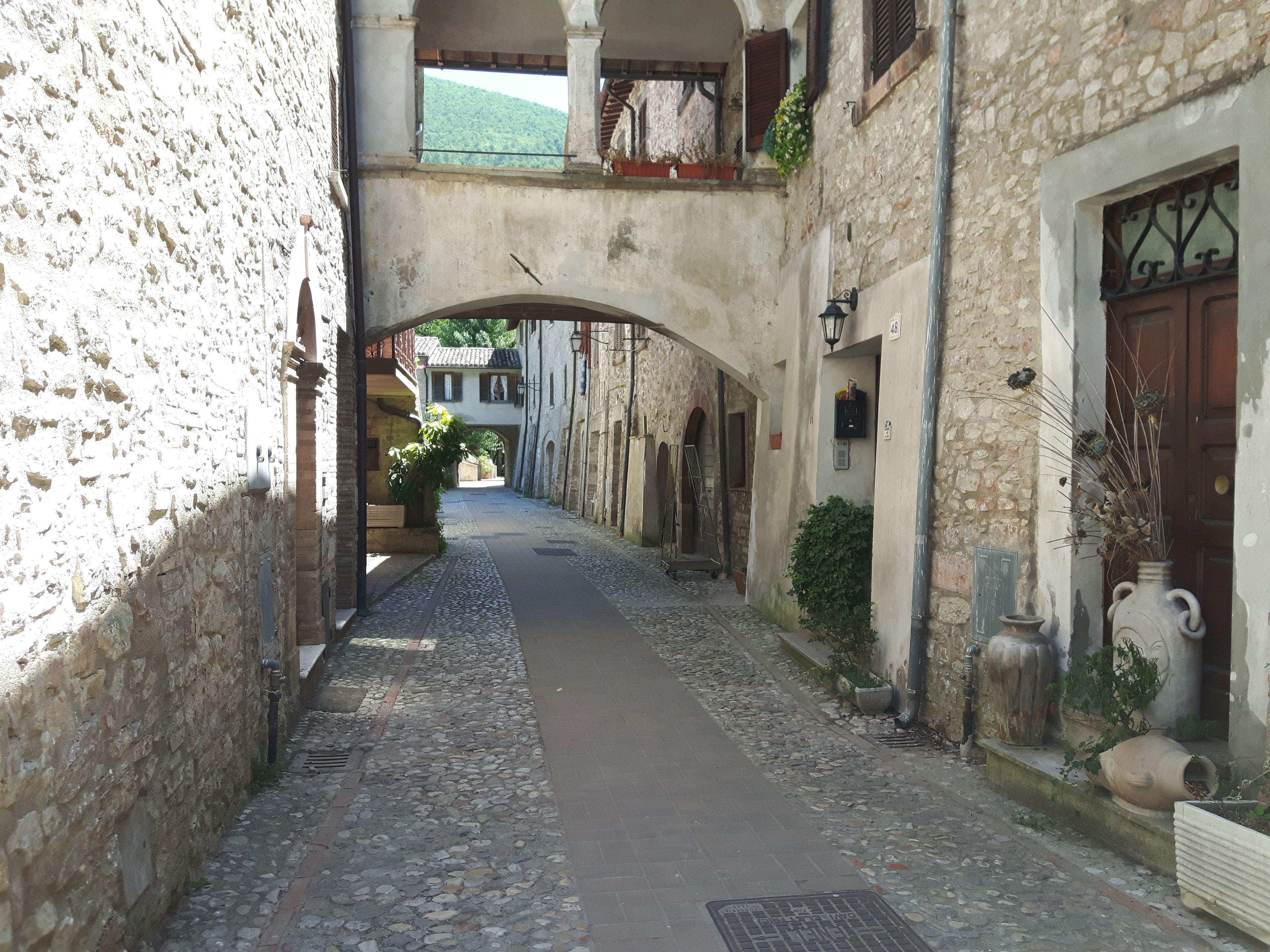 Une ruelle étroite pavée, bordée de bâtiments en pierre dotés de portes et de fenêtres en arc de cercle, qui mène à un passage couvert.