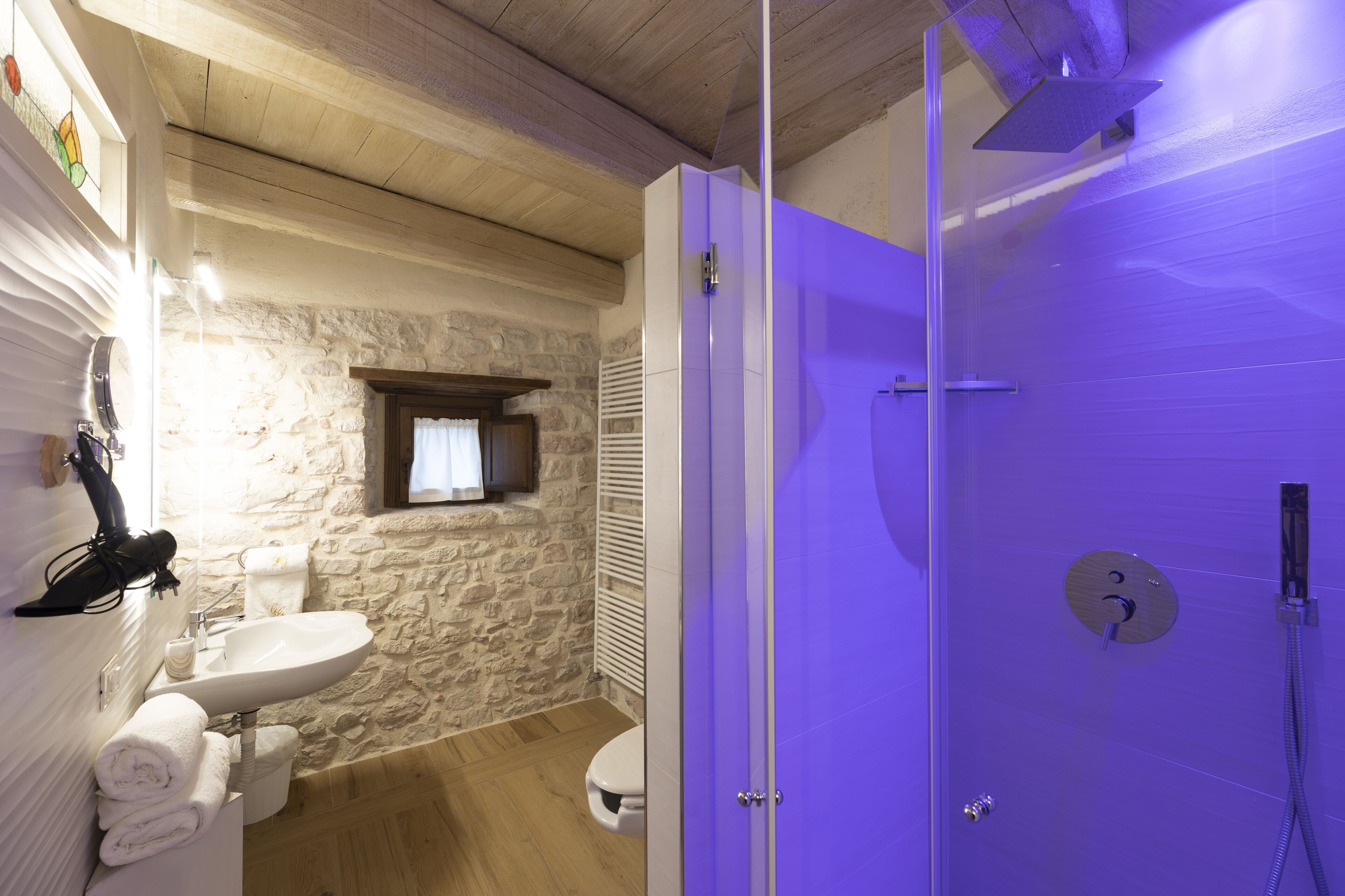 Un baño con pared de piedra, suelo de madera, lavabo blanco y cabina de ducha morada.
