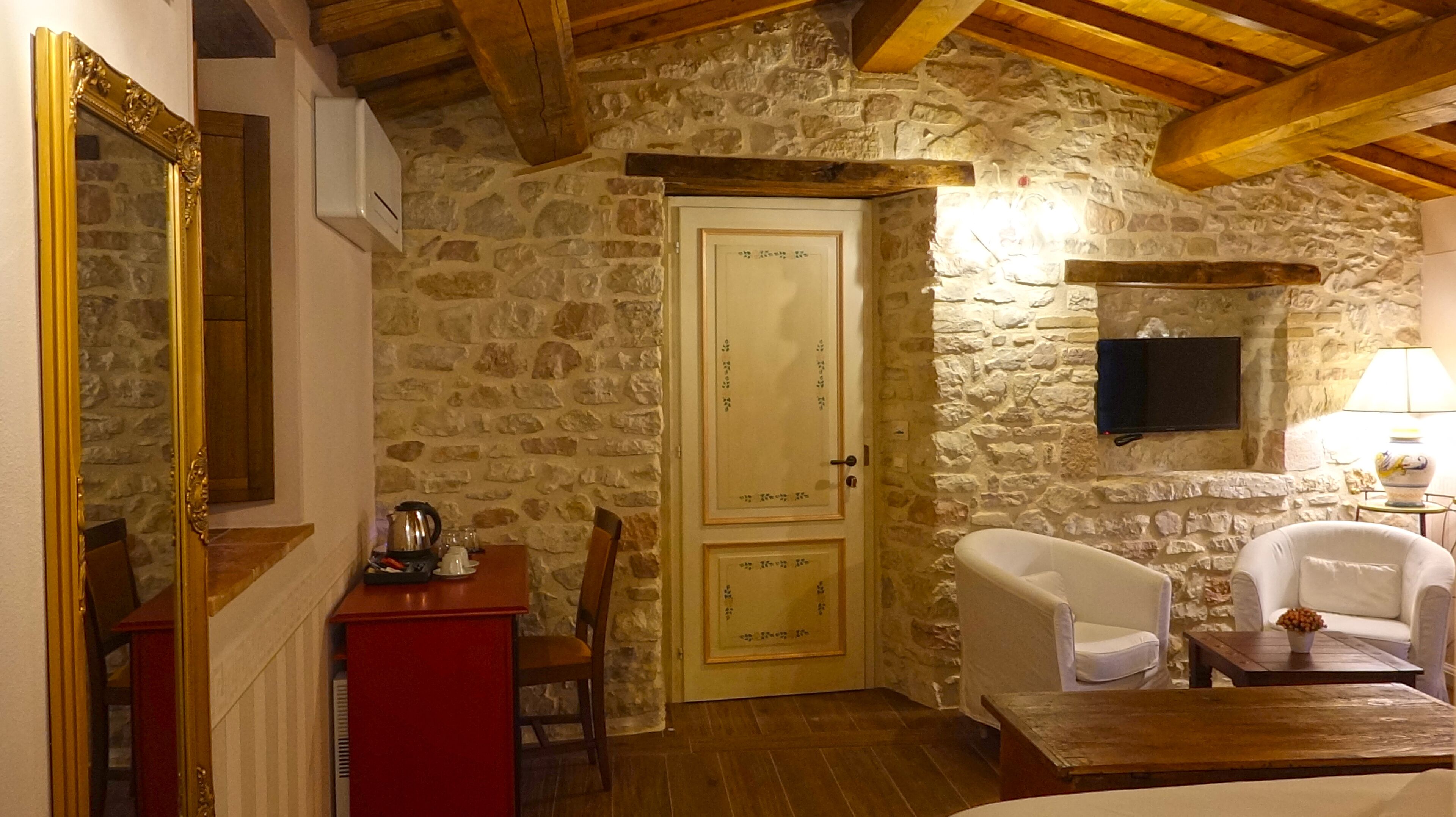 A stone-walled room with a wooden ceiling, a door, a television mounted on the wall, a chair, a small table, and a red cabinet.