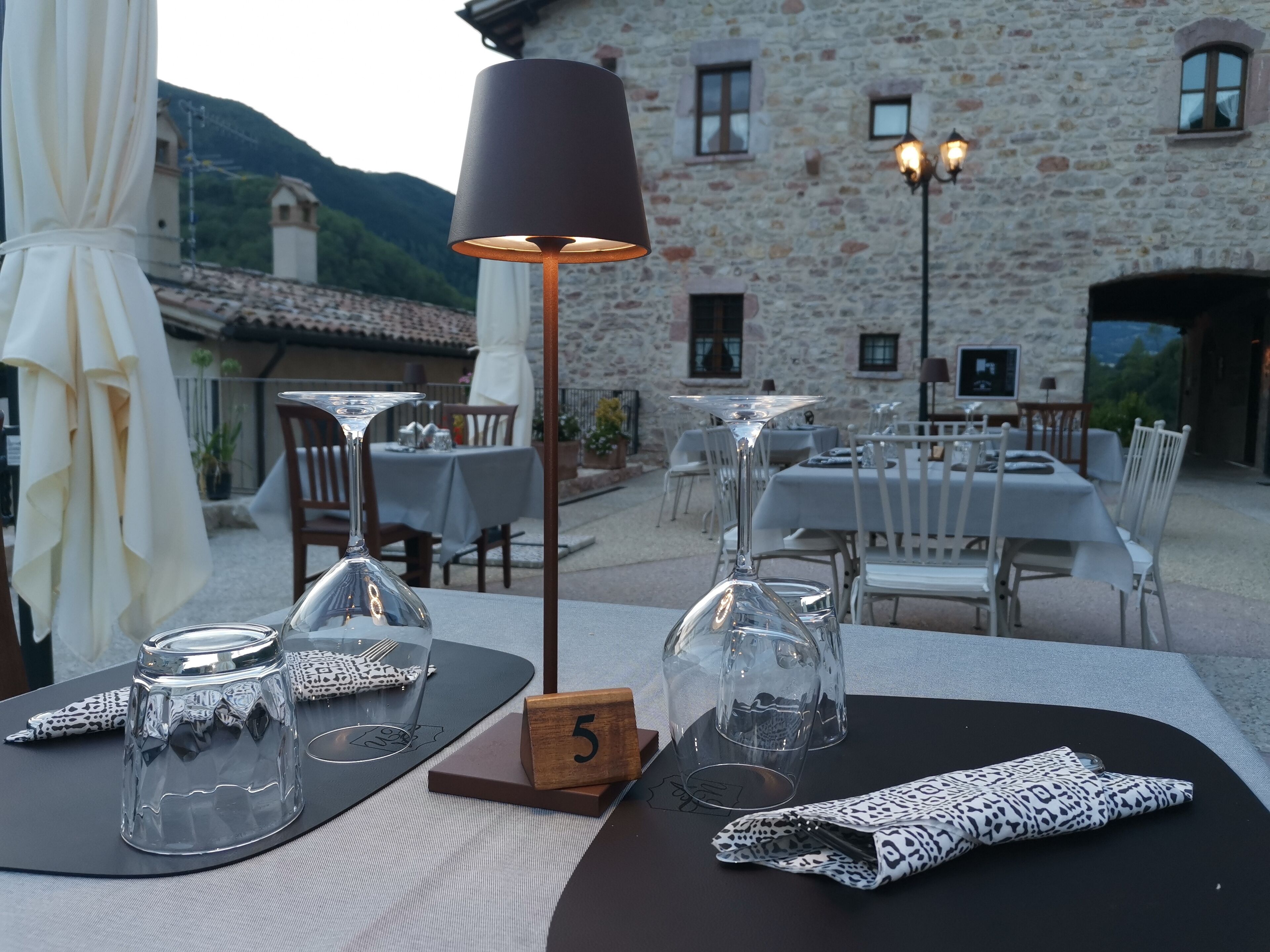 Un área de comedor al aire libre con mesas preparadas para dos personas, una lámpara y un bloque con el número 5.