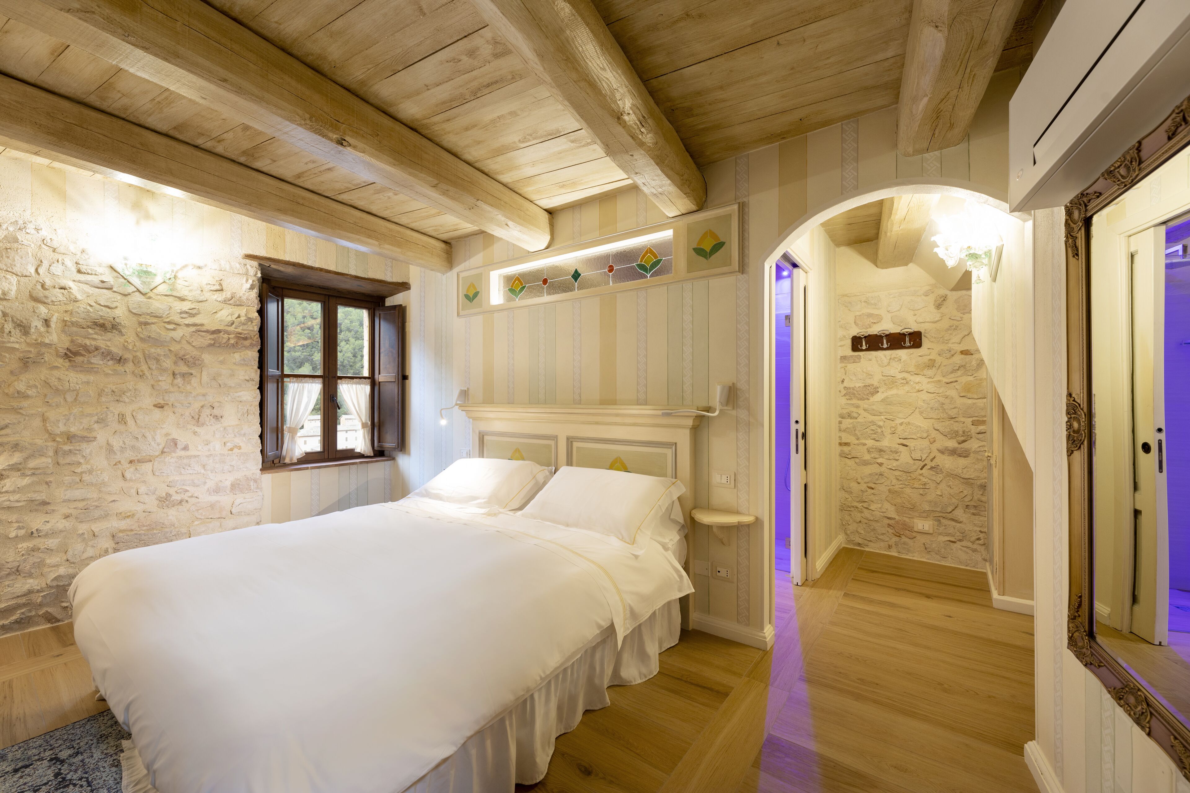 A bedroom with a large bed, wooden ceiling beams, stone walls, and a small window.