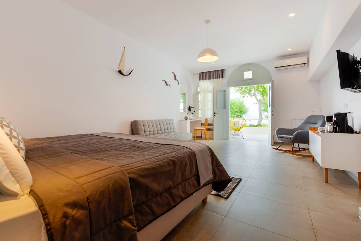 Une chambre moderne avec un grand lit, un bureau et une chaise. On peut admirer la vue sur l’extérieur à travers une porte ouverte.