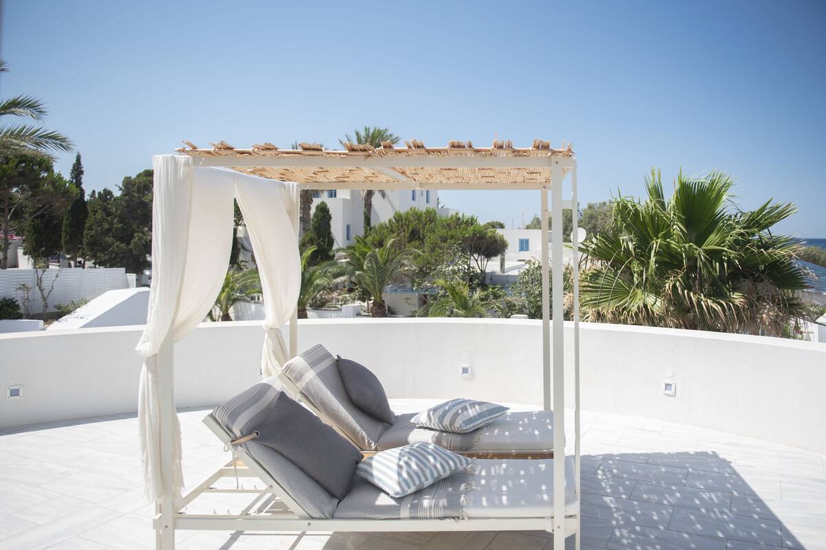 Un espace extérieur aménagé avec des sièges blancs, doté d’un auvent, de fauteuils confortables et offrant une vue sur la mer et les bâtiments en arrière-plan.