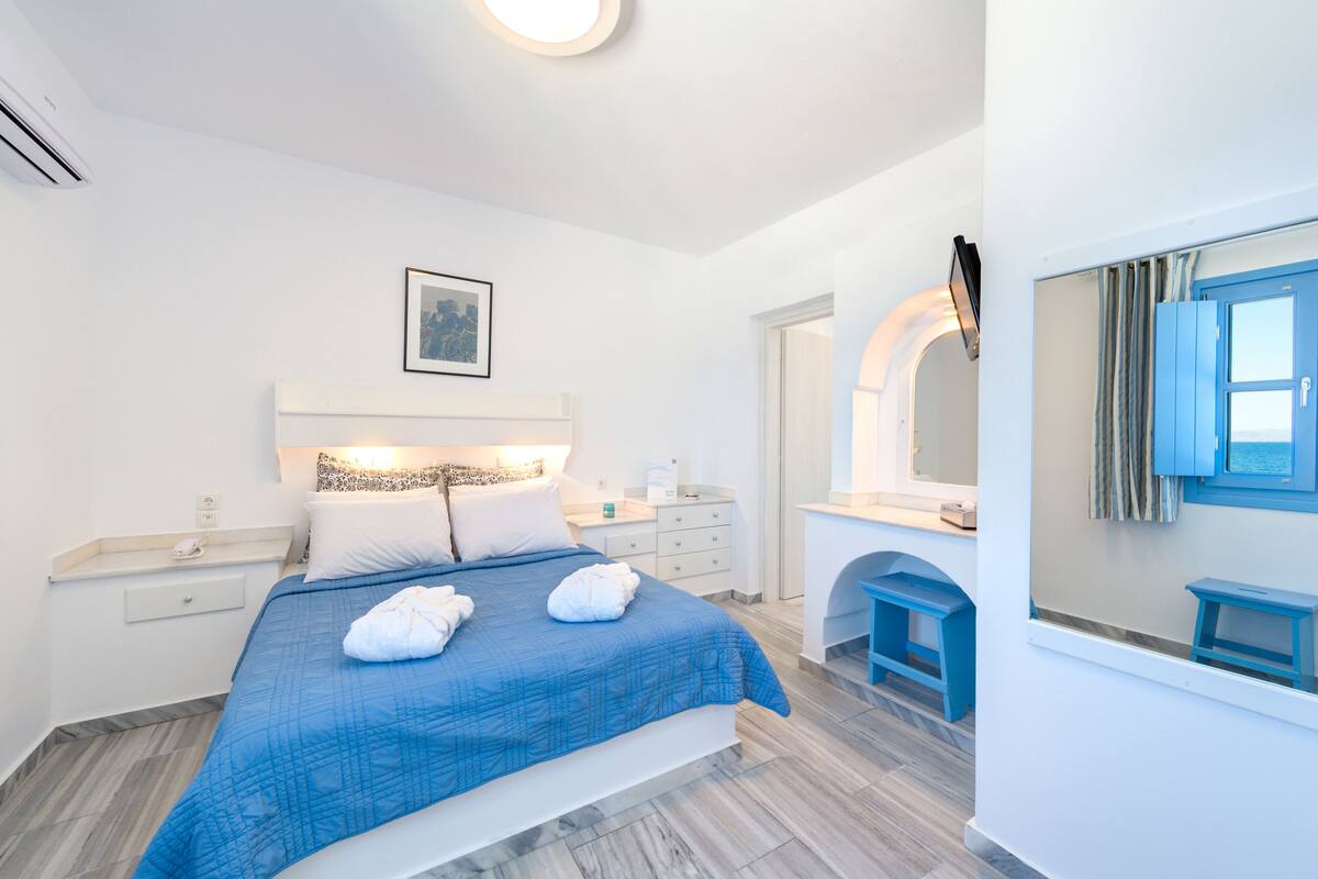 Une chambre d’hôtel moderne avec un grand lit, une table de chevet, un miroir et une salle de bain dotée d’un lavabo et d’une douche bleus.
