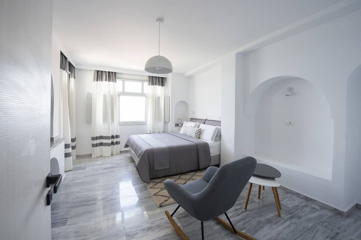 Une chambre moderne avec un lit, un fauteuil gris, une petite table et une grande fenêtre avec des rideaux.