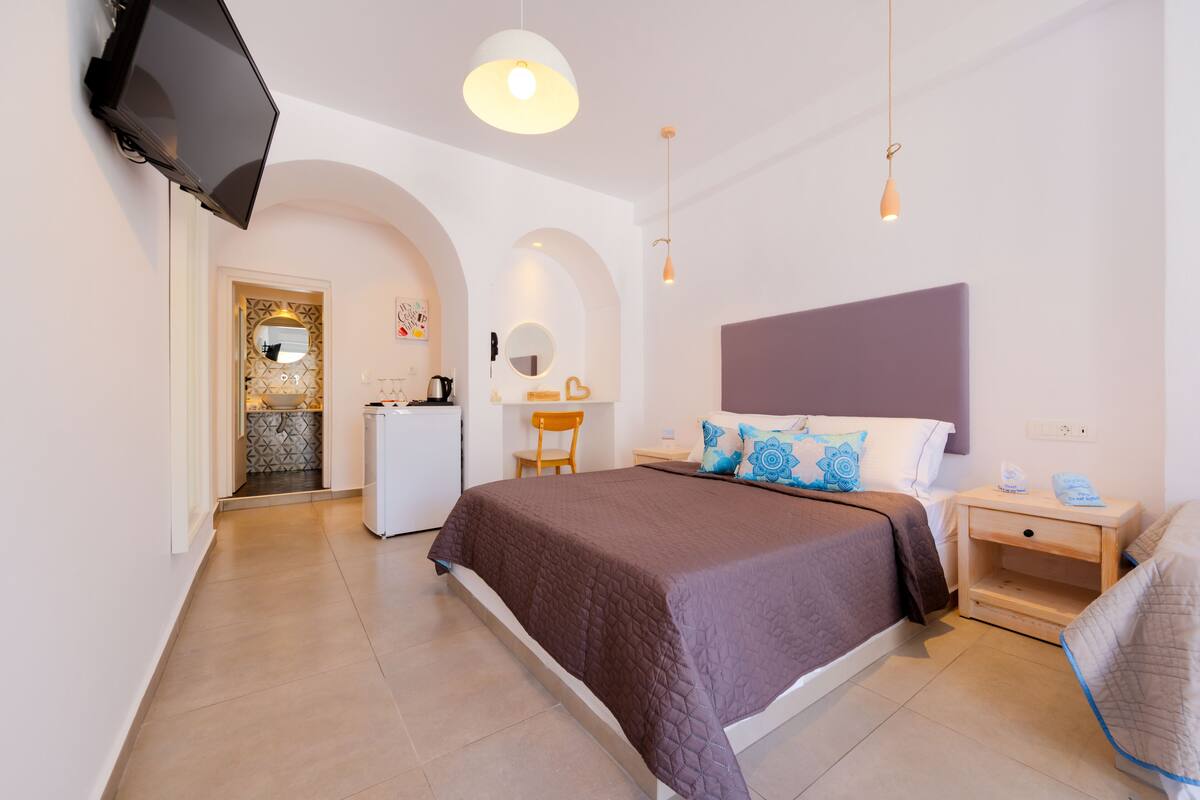 Une chambre d’hôtel moderne avec un grand lit, un téléviseur fixé au mur, un bureau avec une chaise et, au fond, une petite kitchenette.