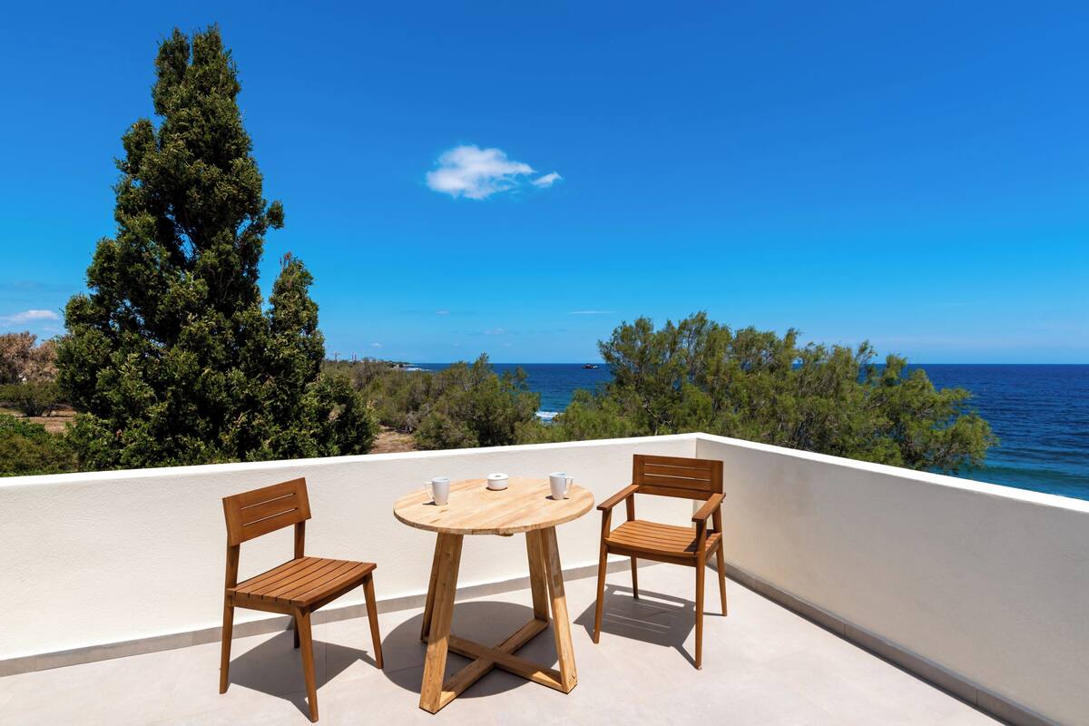 Une table et des chaises en bois sur un balcon donnant sur la mer et les arbres.