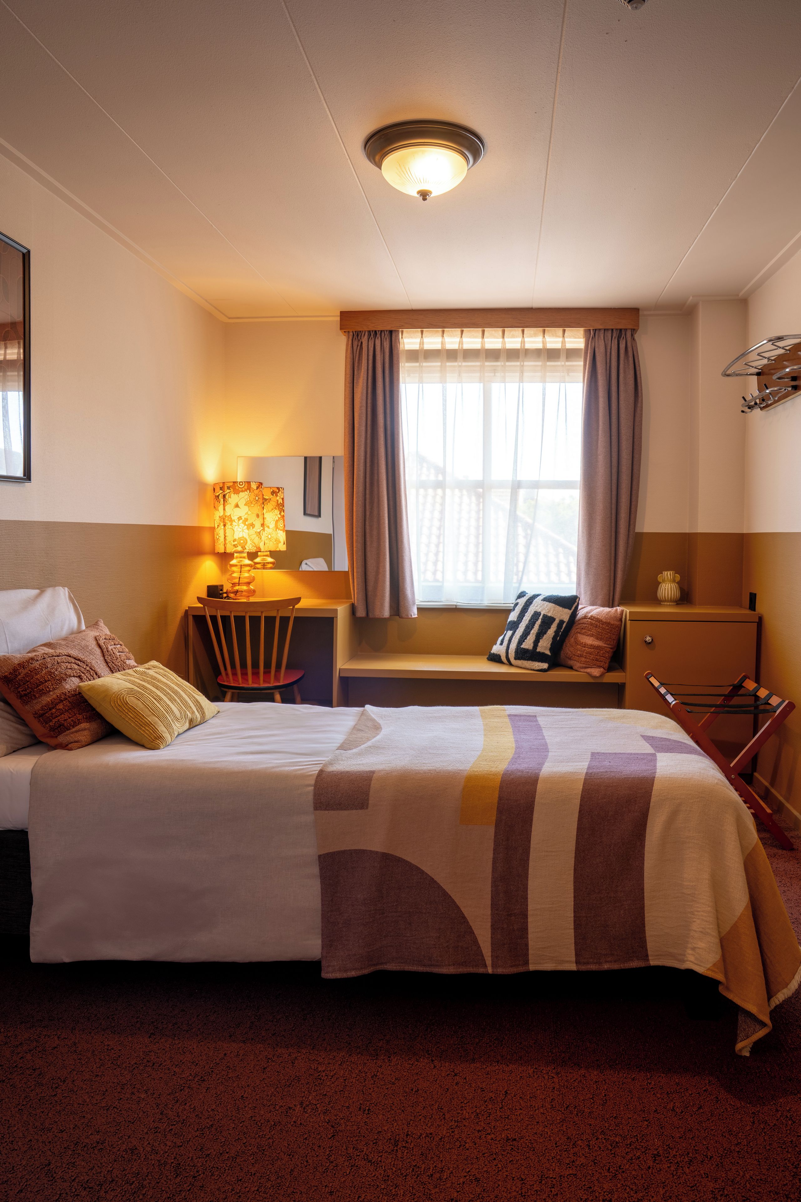 Une chambre d’hôtel avec un lit, un bureau, une chaise, une fenêtre avec des rideaux et une lampe.