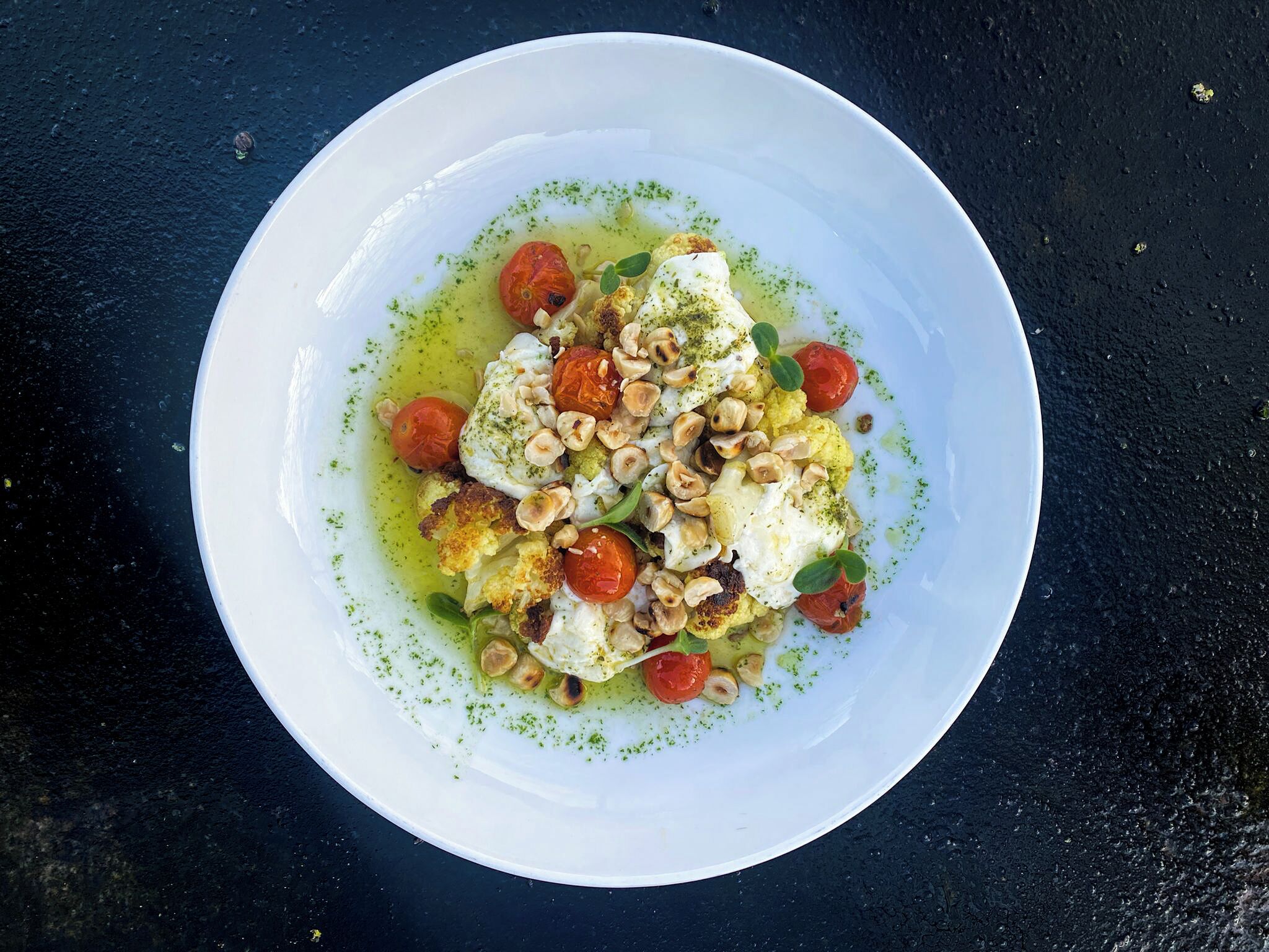 A white plate with a dish of poached eggs, cherry tomatoes, and roasted nuts on a dark surface.
