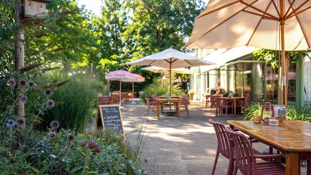 An outdoor patio with wooden tables and chairs, surrounded by lush greenery and large umbrellas. - Hotel Restaurant Ruimzicht (Zeddam)