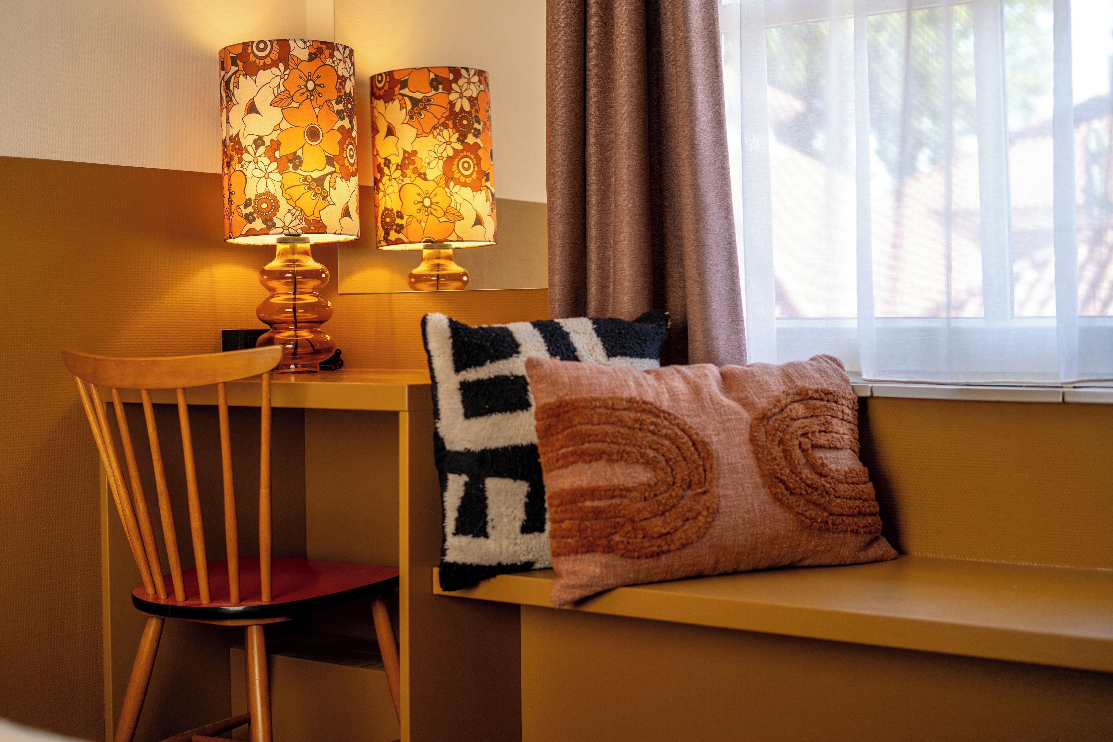 A room with a wooden chair, a bench with two pillows, and two lamps with floral patterns.