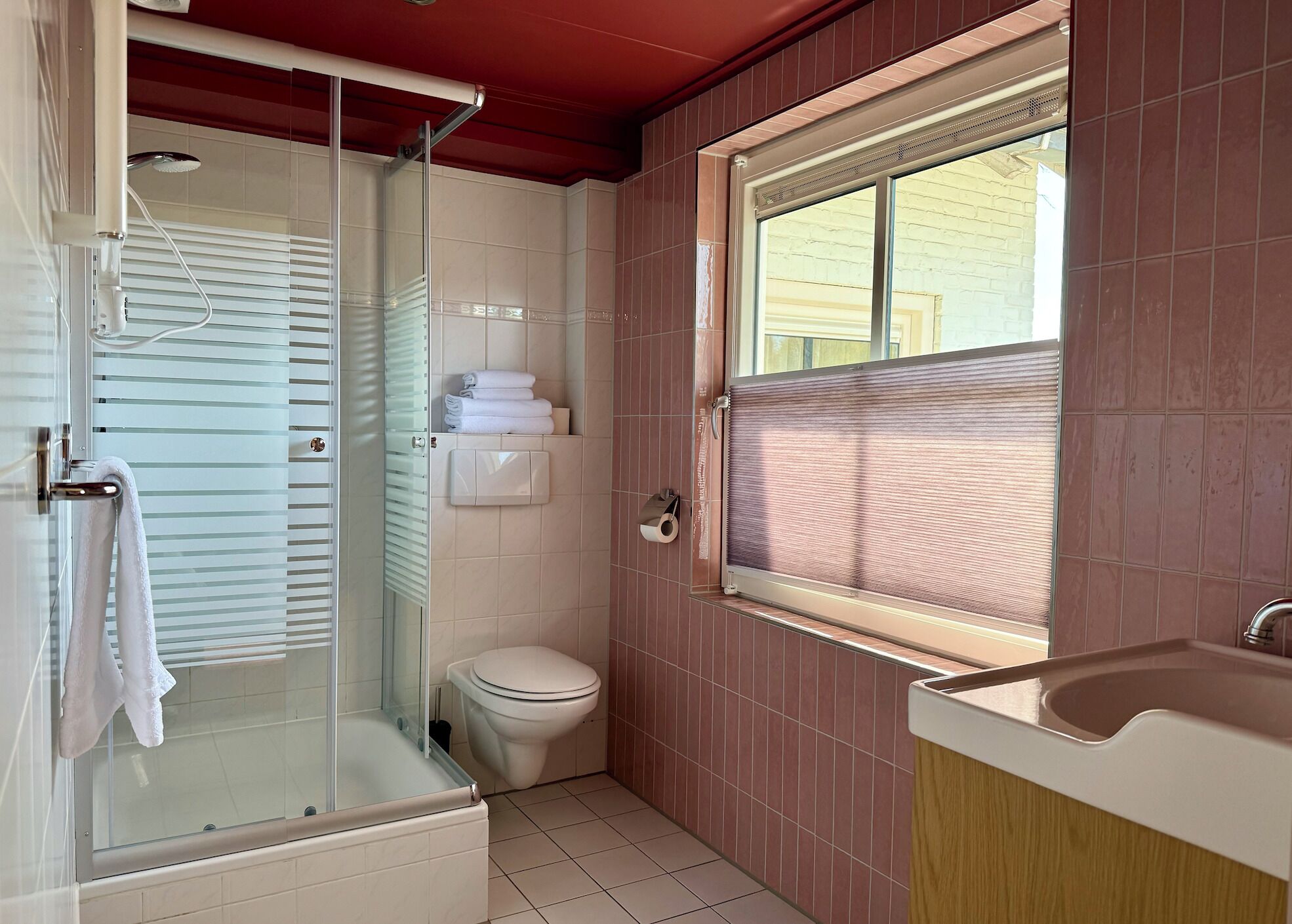 A bathroom with a glass-enclosed shower, a toilet, a sink, and a window with blinds.