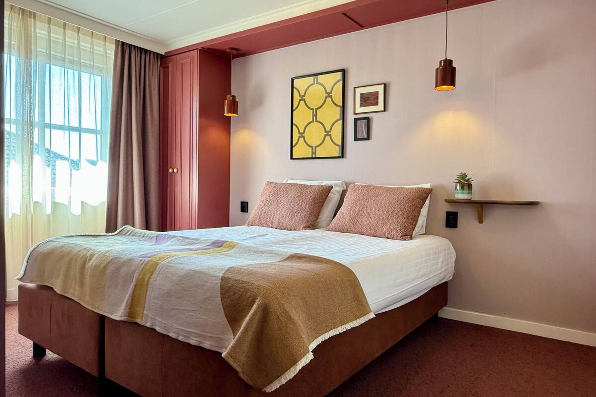 A neatly made bed with a patterned bedspread, a bedside table with a plant, and a wall-mounted shelf.