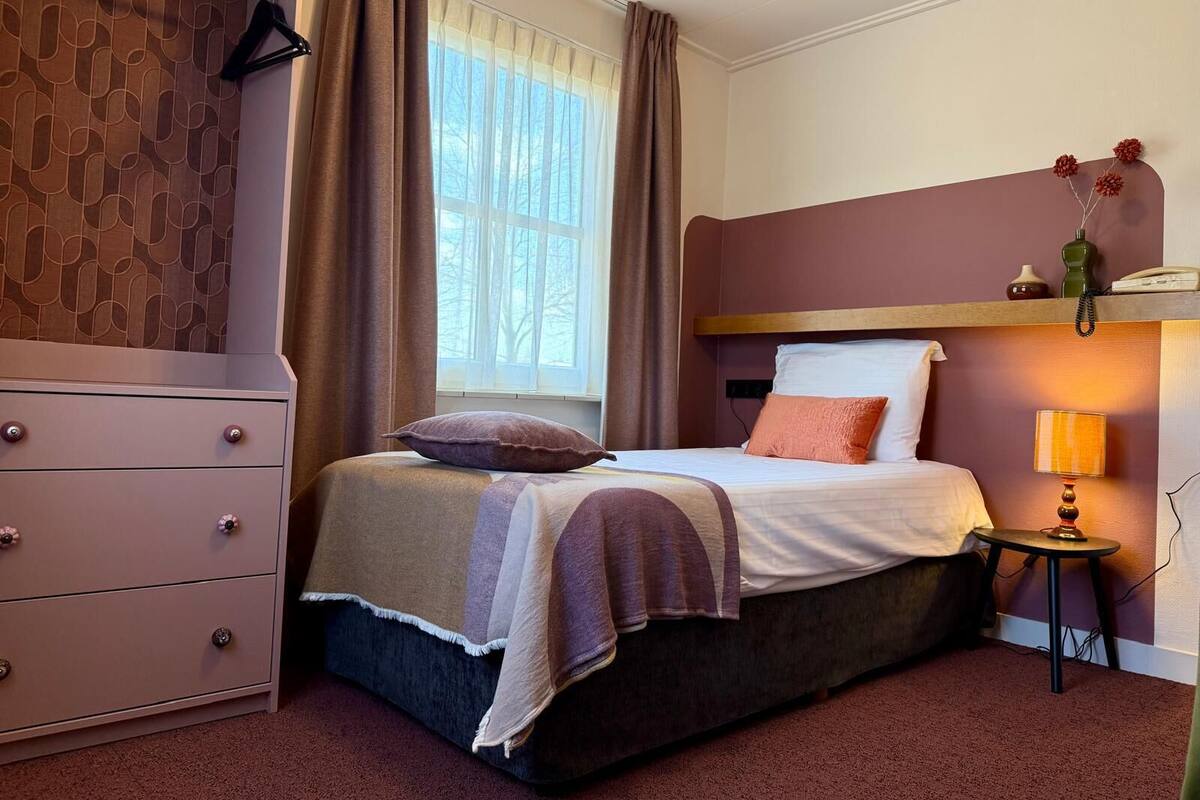 A bedroom with a bed, a dresser, a window with curtains, a bedside lamp, and a small table with a vase.