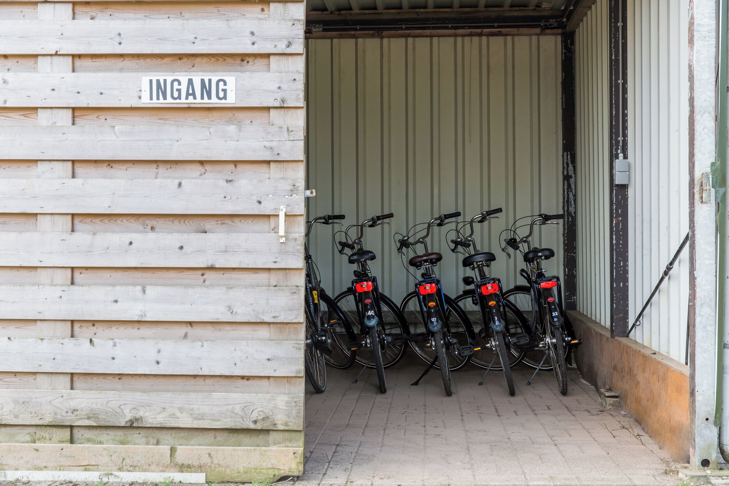 A wooden building entrance with a sign reading "INGANG" and a row of bicycles parked inside.