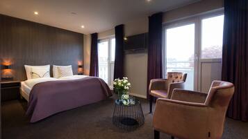 Een moderne hotelkamer met een groot bed, een zithoek met twee stoelen, een klein bijzettafeltje met een bloevas en een groot raam met uitzicht.