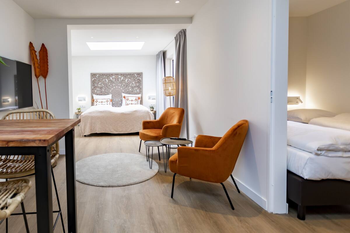 A modern hotel room with a bed, two orange armchairs, a small table, a TV, and a large window with curtains.