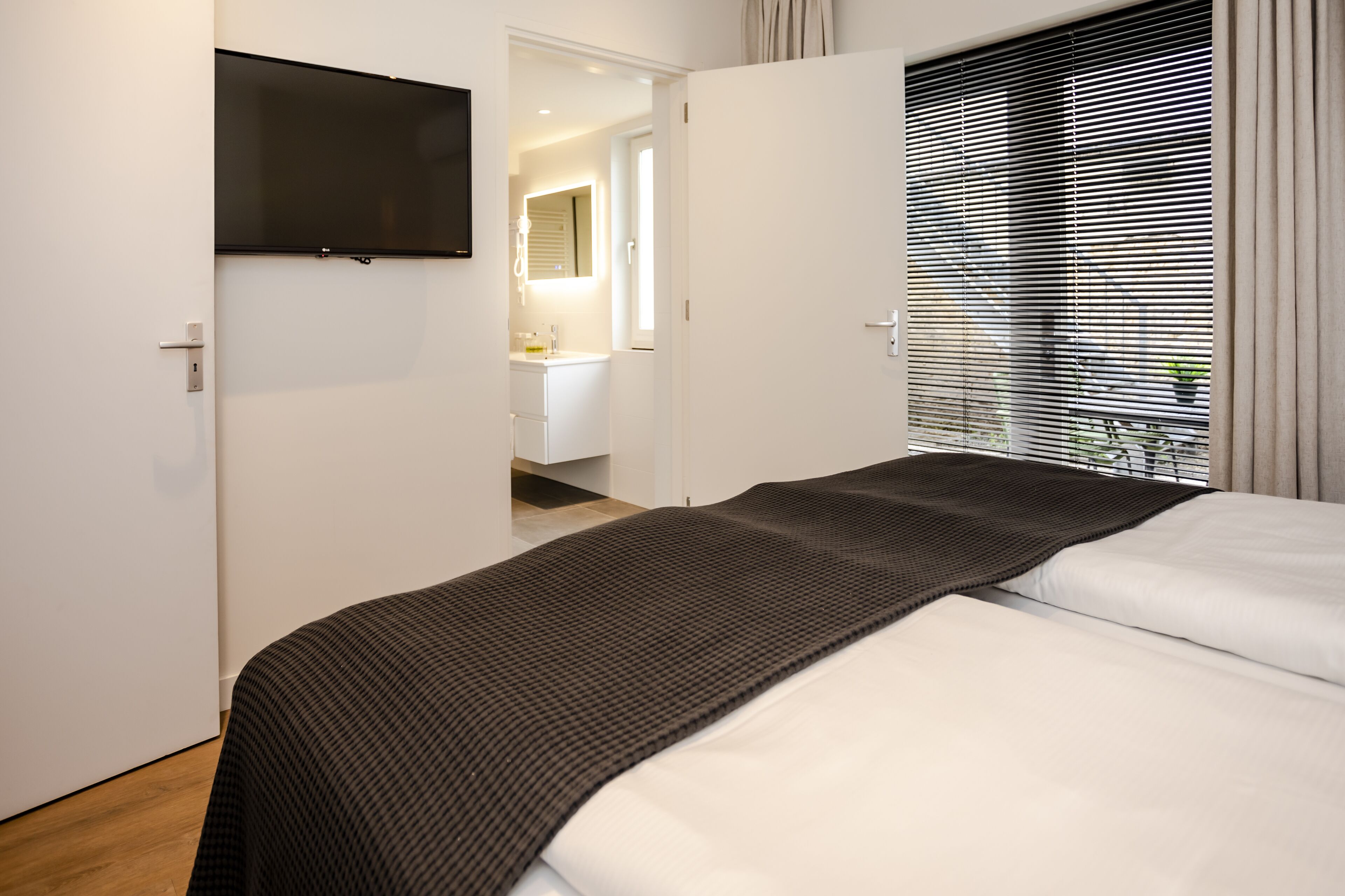 Et moderne hotelværelse med en seng, et fjernsyn ophængt på væggen, et badeværelse, der kan ses gennem en åben dør, og et vindue med persienner.