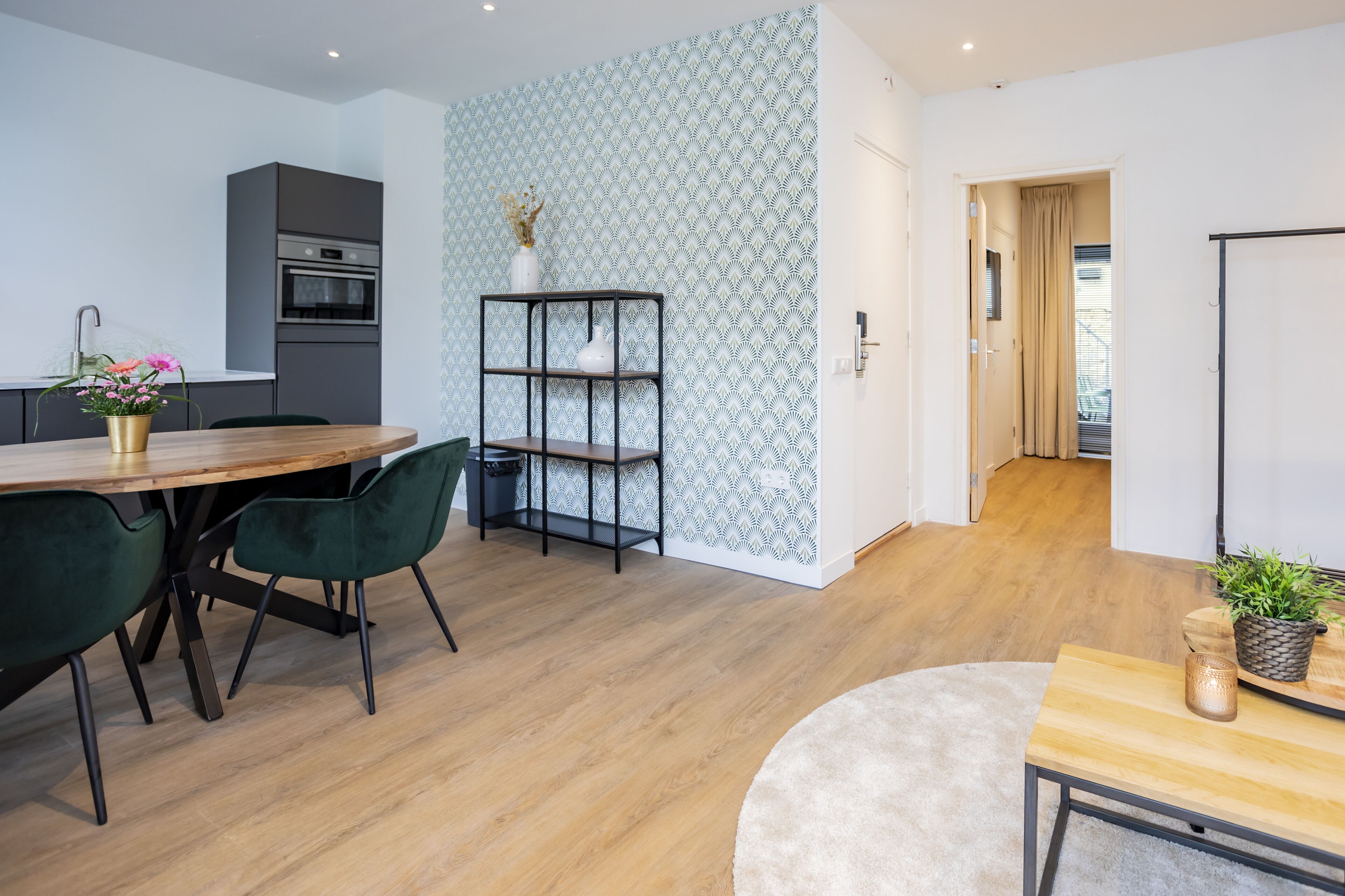 A modern kitchen with a dining area, featuring a wooden table, green chairs, a black metal shelf, and a patterned wall.