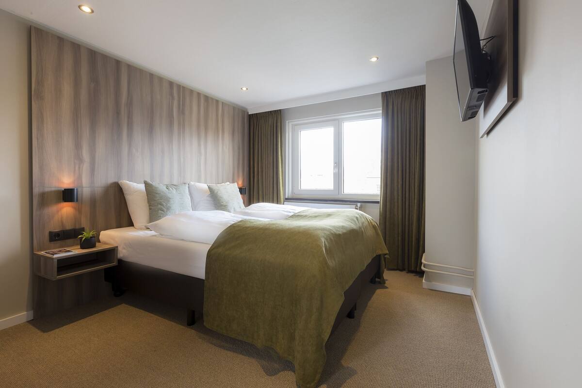 A modern hotel room with a large bed, a bedside table with a lamp, a flat-screen TV mounted on the wall, and a window with curtains.